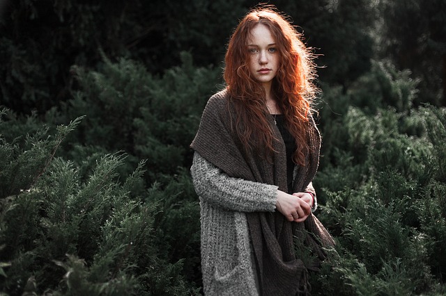Dievča s dlhými ryšavými vlasmi, v sivom svetri, so šálom stojí pred lesom